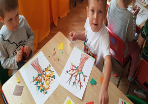 dzieci przy stoliku robią wydzierankę z kolorowego papieru (liście) i naklejają na kontur drzewa